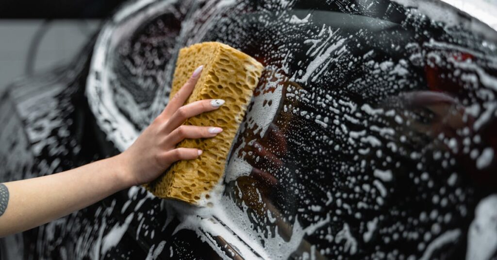 A hand scrubs a car window with a sponge, covered in soap and foam, at a car wash.